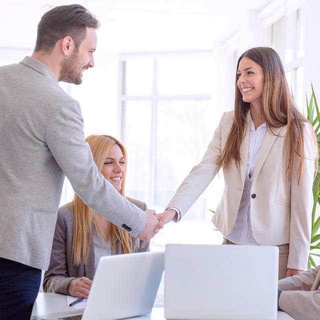 Vier Personen in einem Bürogebäude, zwei stehen und geben sich die Hand, zwei sitzen am Tisch und beobachten Vier Personen in einem Bürogebäude, zwei stehen und geben sich die Hand, zwei sitzen am Tisch und beobachten