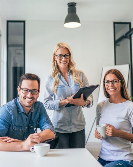 Drei Personen sitzen an einem Tisch in einem modernen Büro mit großen Fenstern und arbeiten gemeinsam an einem Laptop Drei Personen sitzen an einem Tisch in einem modernen Büro mit großen Fenstern und arbeiten gemeinsam an einem Laptop