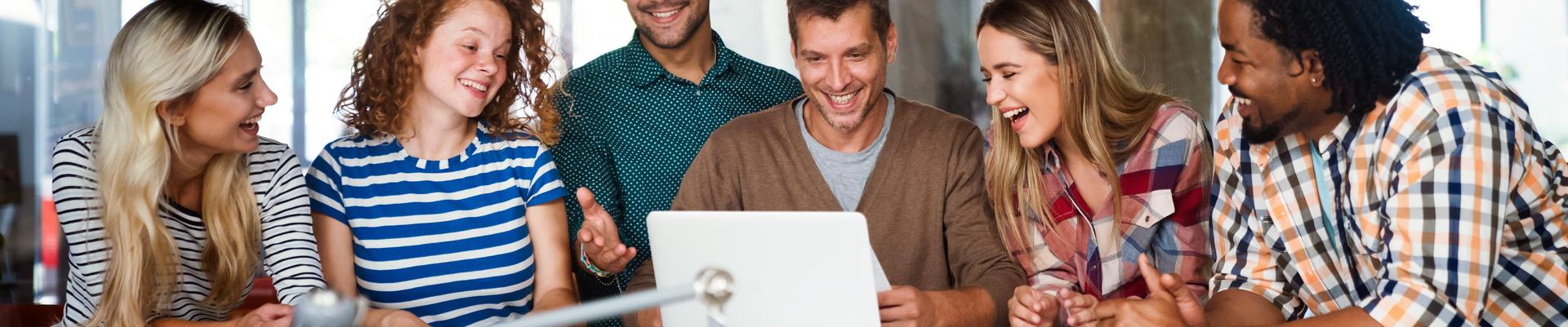 Sechs junge Erwachsene sitzen um einen Tisch und schauen gemeinsam lachend auf einen Laptop in einem modernen Büro