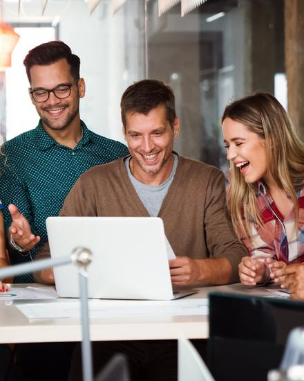 Sechs junge Erwachsene sitzen um einen Tisch und schauen gemeinsam lachend auf einen Laptop in einem modernen Büro