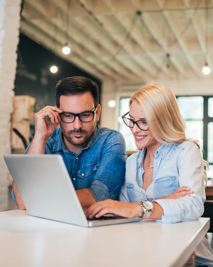 Zwei Personen sitzen an einem Tisch und schauen gemeinsam auf einen Laptop in einem hellen, modernen Büro mit großen Fenstern Zwei Personen sitzen an einem Tisch und schauen gemeinsam auf einen Laptop in einem hellen, modernen Büro mit großen Fenstern